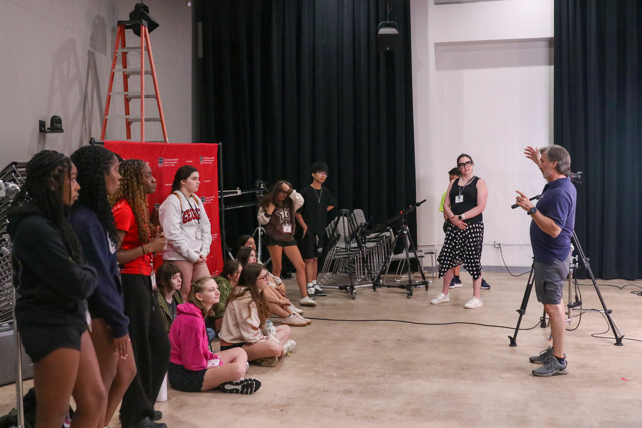 A group of students listens to an instructor gesturing while standing in a studio with black curtains, a camera on a tripod, and a red ladder in the background. Some students are seated on the floor, others stand.