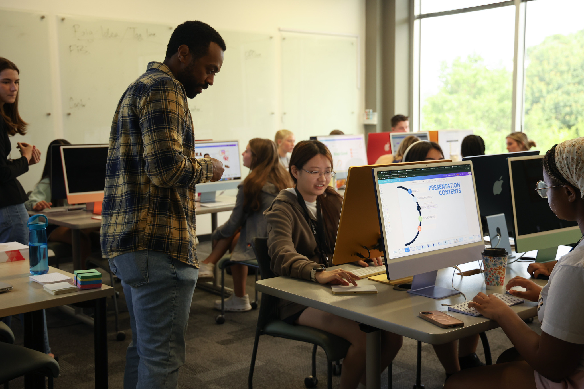 A classroom with students at computers, focused on work. A teacher stands beside a student, assisting her. Large monitors, notebooks, and cups are on the desks. A presentation titled Presentation Contents is open on one screen.
