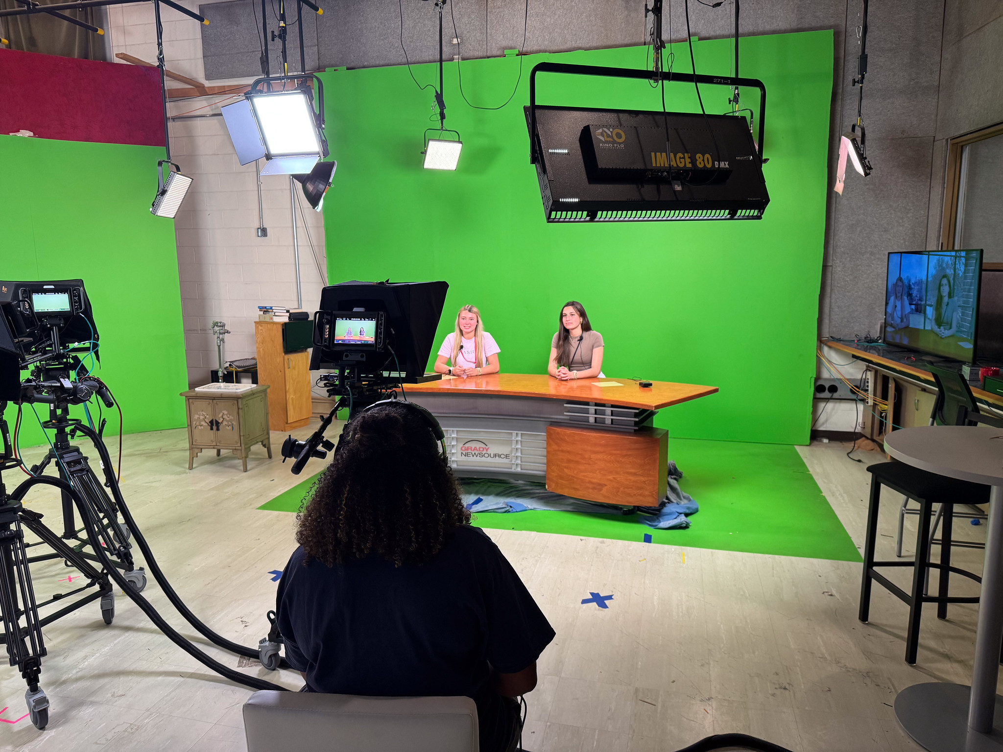 Two people sit at a news desk in a studio with a green screen behind them. Cameras, lights, and crew are visible. A TV monitor shows the live feed. The setting appears to be a news or talk show recording.