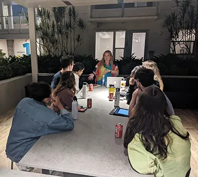 Kathryn Kostovetsky stands at the head of the table talking as a group of eight students listen.