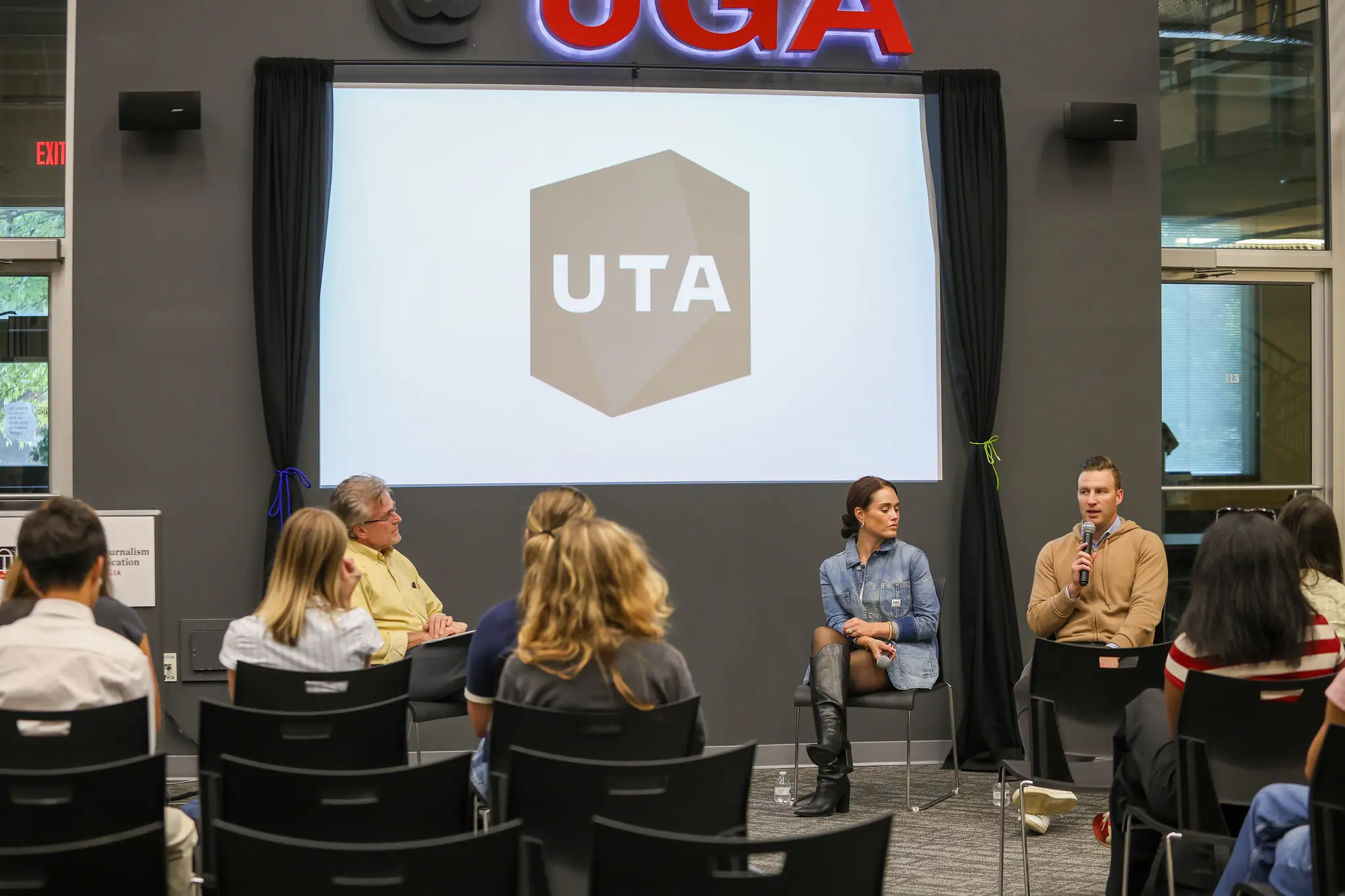 A panel discussion with two professionals in front of a group of students. In the background is a digital screen reading UTA.