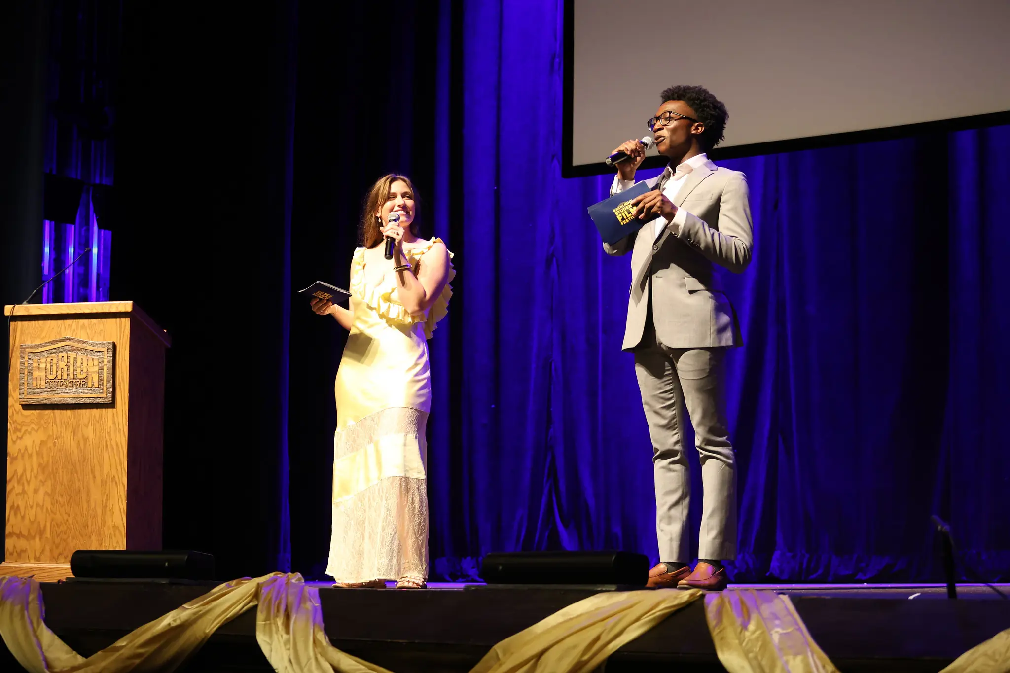 Two students in formal attire address a group from a stage.