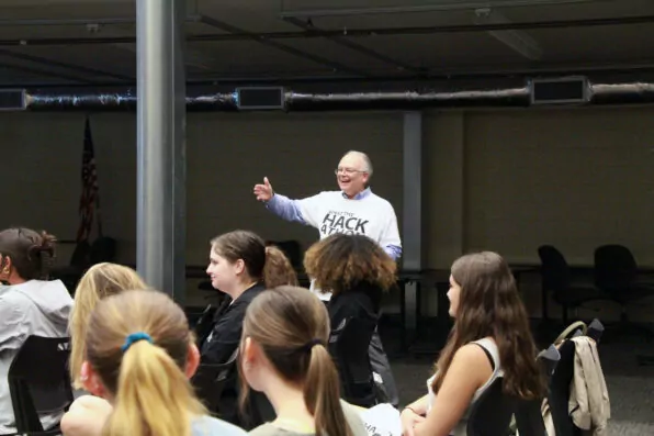 A smiling older adult wearing glasses and a Hack 414 shirt stands among seated students, gesturing with a thumbs up in a classroom or seminar setting. An American flag is visible in the background.