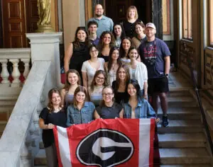 A large group of students and professors stand on stairs and the front row is holding a UGA flag.
