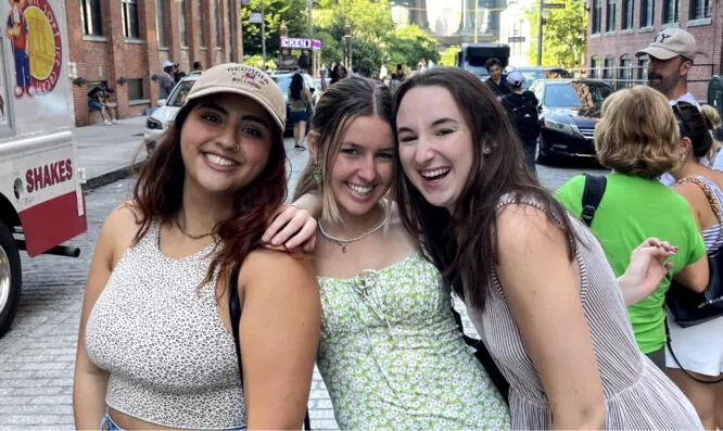 Three young women stand close together, smiling at the camera on a sunny cobblestone street, enjoying a break during their NYC internship. People and cars bustle in the background.