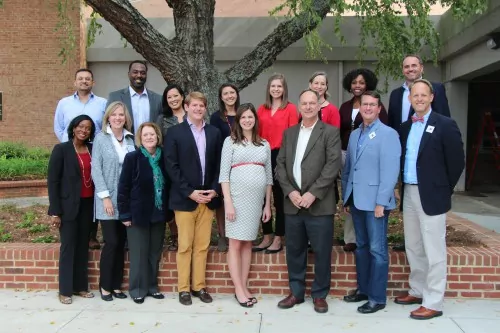A group of fifteen adults pose and smile for a photo outdoors in front of a large tree and brick building. They are dressed in business casual attire and stand in two rows on a sidewalk.