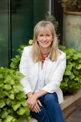 A woman with blonde hair, wearing a white blazer, white blouse, and blue jeans, sits outdoors on a low wall surrounded by green plants, smiling at the camera.
