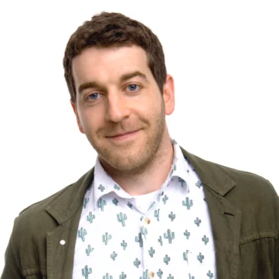 A man with short brown hair and blue eyes smiles slightly. He is wearing a white shirt with a green cactus pattern and a green jacket. The background is plain white.
