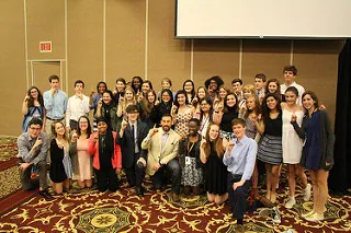 A large group of young adults and a few older individuals pose together indoors on a patterned carpet, dressed in semi-formal attire, smiling and making hand gestures toward the camera.