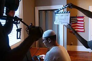 A person wearing a white cap sits at a desk while a film crew prepares to shoot, with a clapperboard and camera visible. An American flag hangs on the wall in the background.