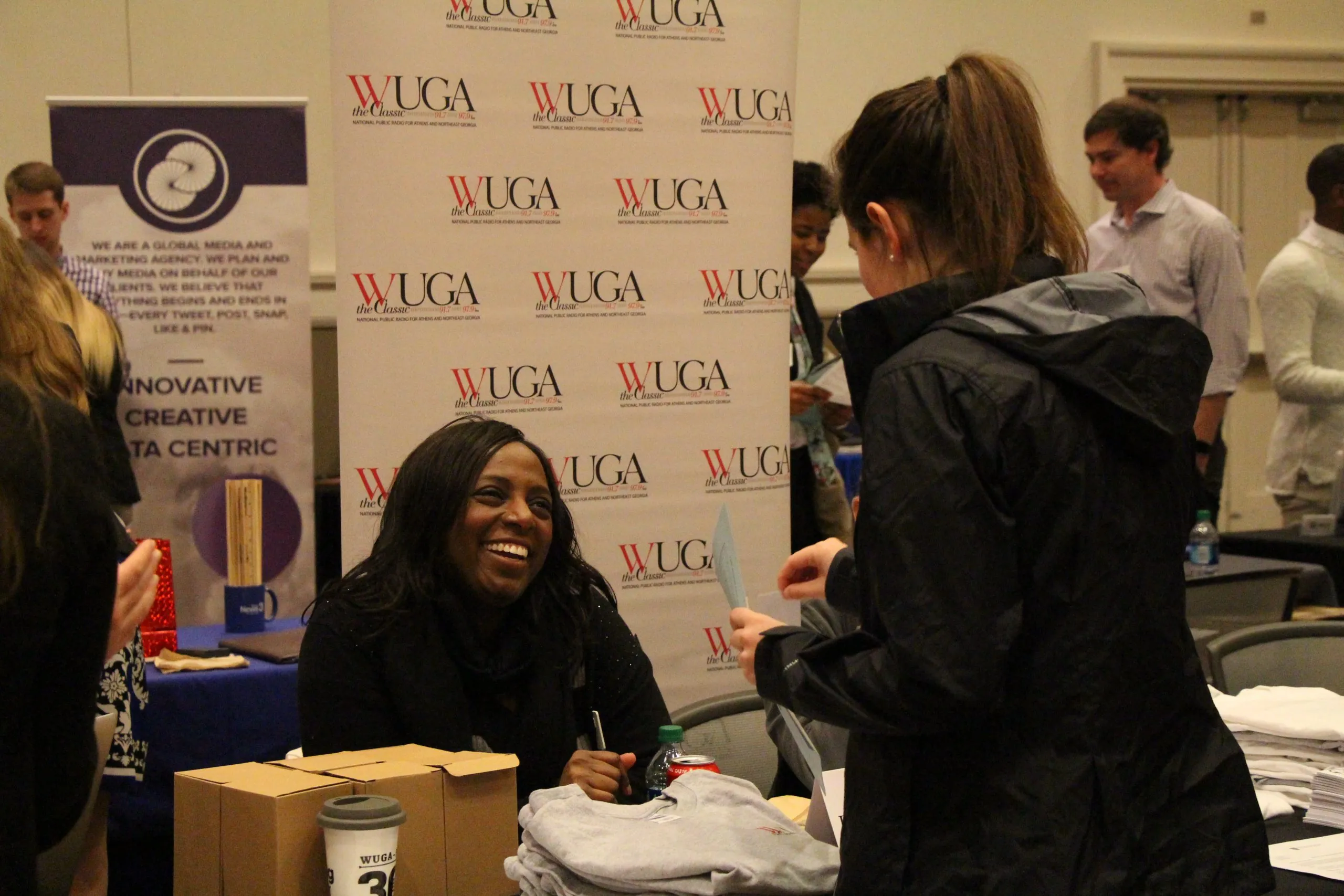 A woman sitting at a table smiles and talks with a standing visitor at an event. Promotional banners and tables with t-shirts, boxes, and other materials are visible in the background.