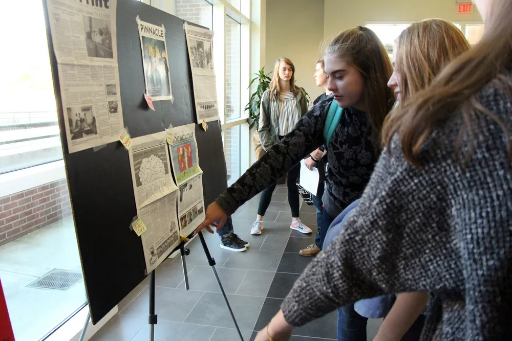 Several students stand by a display board covered with newspaper clippings. One student in the foreground points at the board, while others observe, in a well-lit hallway with large windows.