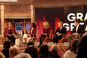 Five people sit on stage in director’s chairs having a panel discussion in front of a live audience. A large screen behind them displays partial text. The setting appears to be a talk show or a public event.
