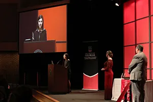 A woman speaks at a podium on stage during an event, with her image projected on a large screen. Two other people stand nearby in front of a red and black Georgia banner and a table with awards.