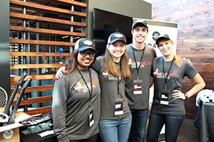 Four smiling people wearing matching gray shirts and hats with Next Media logos stand together indoors. They each have name badges and their arms around each other, posing for a group photo at an event booth.