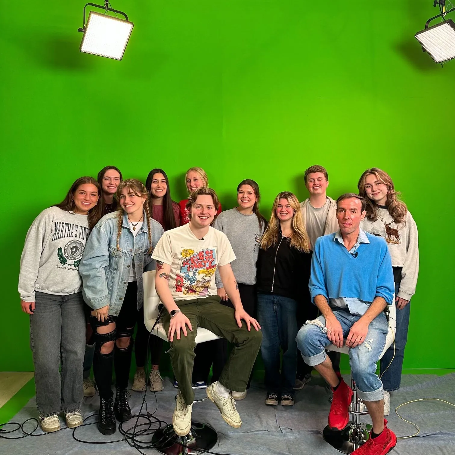 A group of eleven people, including Timi Meade, some standing and two sitting on stools, pose and smile in front of a green screen with studio lights above them. The floor is partly covered with a rug, and cables are visible.
