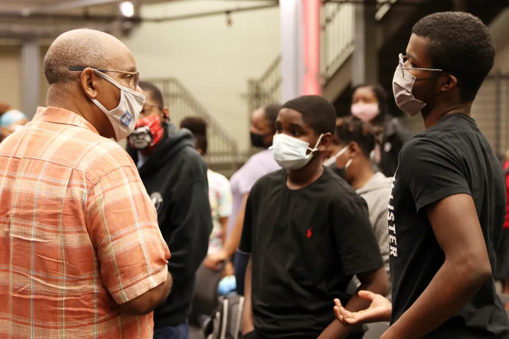 Rick Dunn, in a plaid shirt and glasses, talks with two teenage boys—all wearing face masks—in a busy indoor setting with more people and stairs in the background.