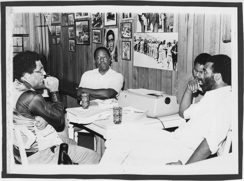 Four men, including Rick Dunn, sit around a table in a wood-paneled room, deep in discussion. The table is scattered with papers, glasses, and a typewriter as framed photos fill the wall behind them.