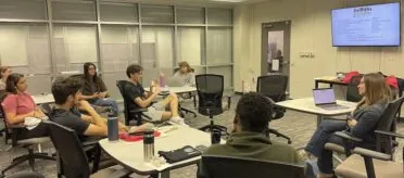 A group of Griffiths Innovators students sit in the JLab with Dr. Amanda Bright in a round-table style discussion to brainstorm ideas for their news outlets.