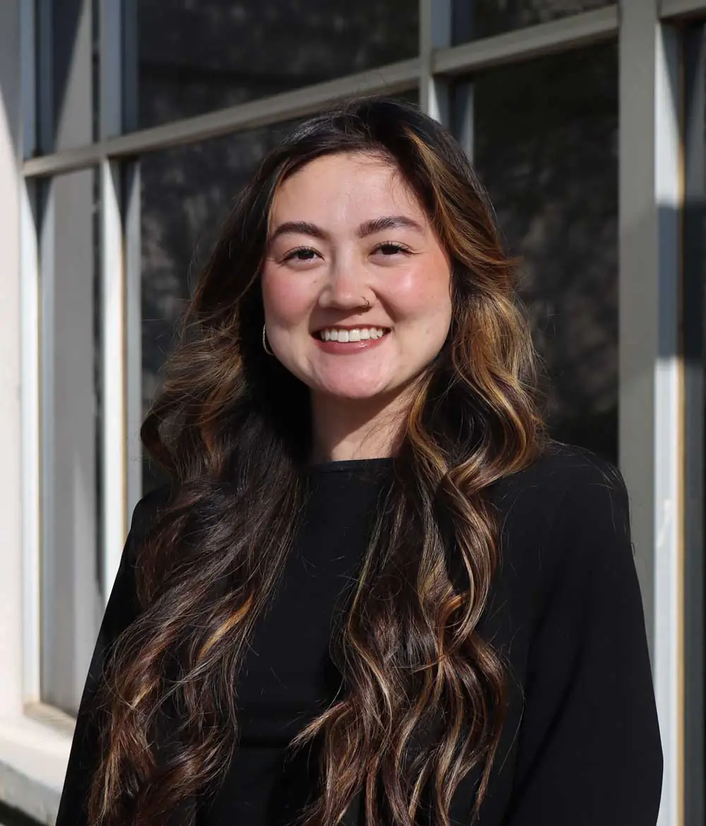 An image of Olivia Ford, a smiling woman in a black shirt with long curled hair in front of a building.