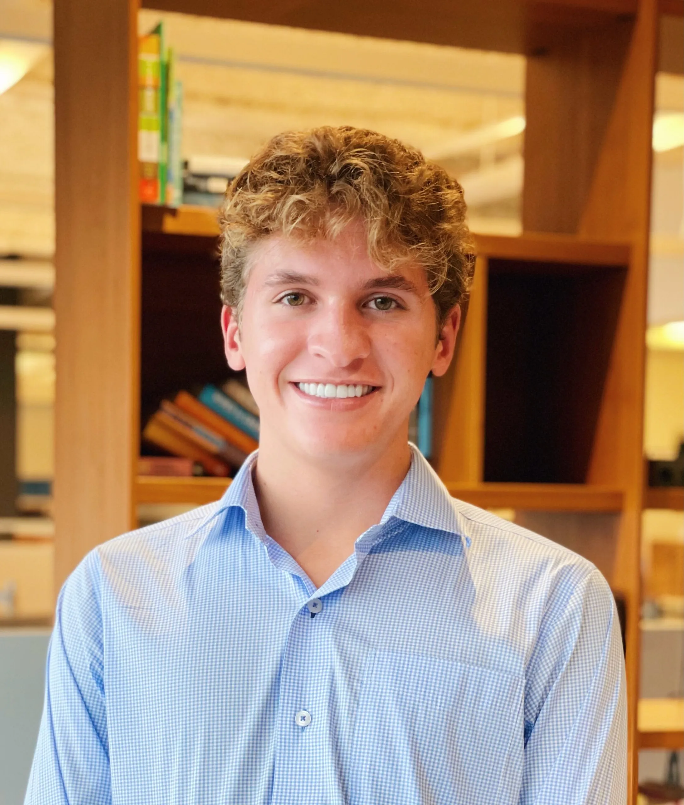 A young man with curly blond hair, wearing a light blue checkered shirt, stands smiling in an office with wooden shelves and books in the background.