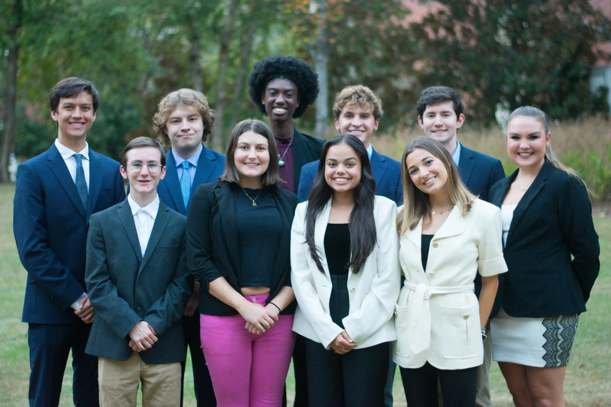 A group of ten young adults, dressed in formal and semi-formal attire, stand together outdoors, smiling at the camera. The background includes grass, trees, and soft-focus greenery.