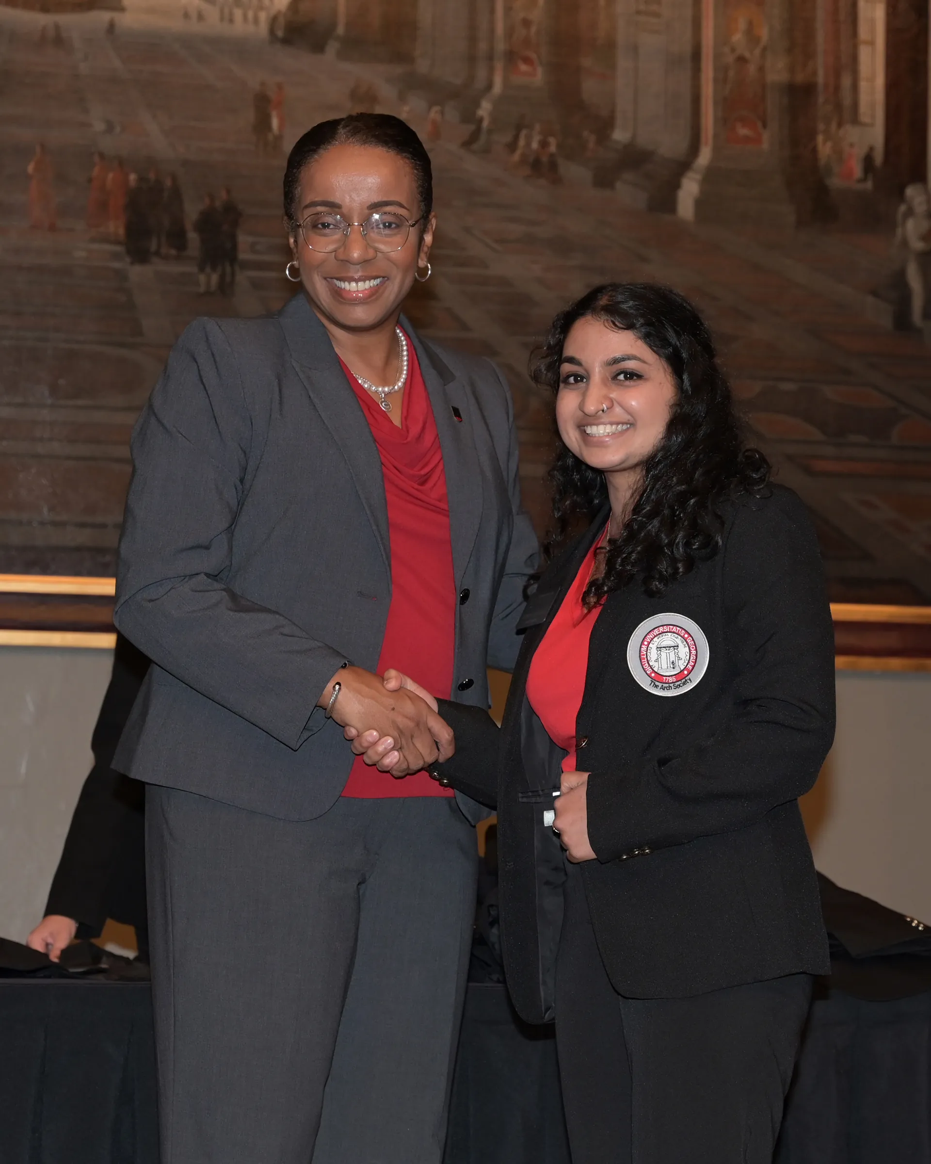 Two women in business attire smile and shake hands at an indoor event. Both wear red tops; one has a suit and glasses, and the other wears a blazer with a circular badge. A large painting is visible in the background.