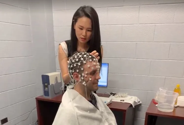 A woman in a lab attaches sensors to a mans head for an EEG test in a clinical setting. The man sits with a towel over his shoulders, while equipment and wires are visible on the table behind them.