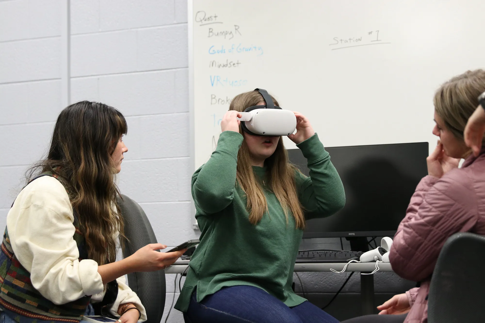 Three women sit in a classroom; one woman in the center is wearing a virtual reality headset, while the others watch and interact. A whiteboard with writing is visible in the background.