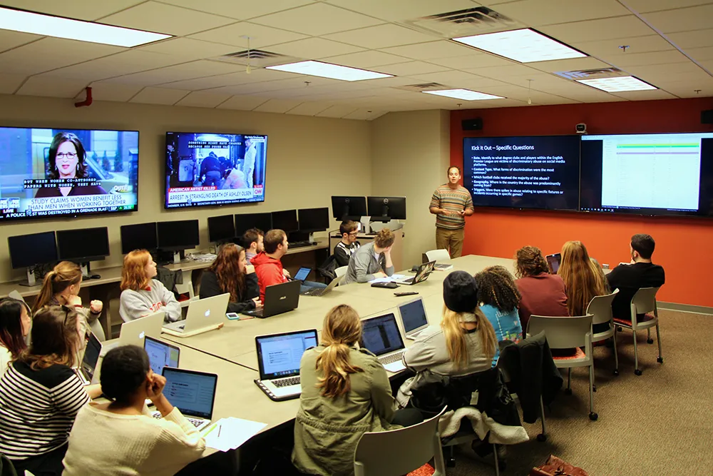 A classroom with students seated around a large table, using laptops while an instructor presents data on a screen. News broadcasts are displayed on two wall-mounted TVs in the background.