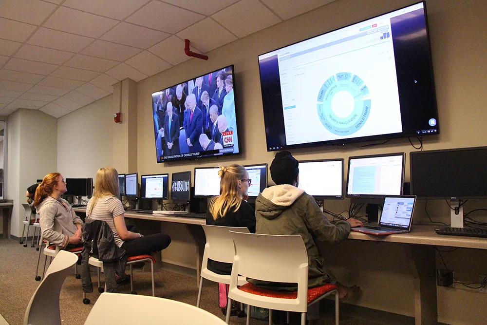 Several people sit at desks using desktop computers in a modern room. Two large screens are on the wall, one showing a news broadcast, and the other displaying a data chart. The atmosphere appears studious and focused.