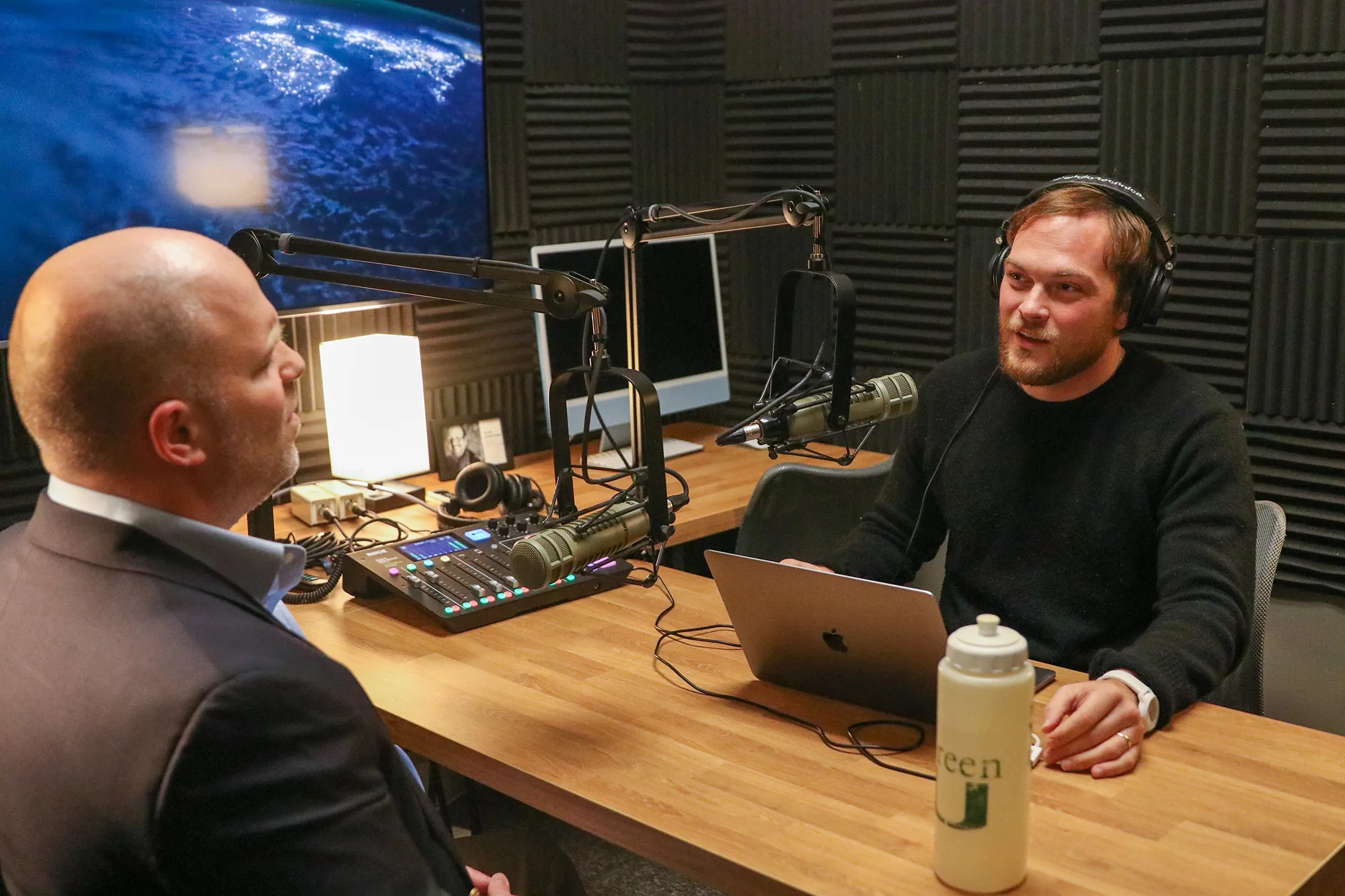 Two men sit at a table in a recording studio with foam wall panels, microphones, and computers. One man speaks into a mic while the other listens. A water bottle and laptop are on the table, with a screen showing Earth in the background.