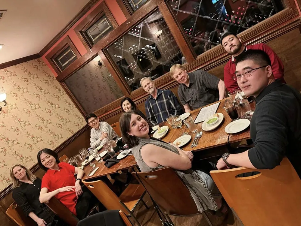 A group of ten people sit around a wooden table at a restaurant, smiling at the camera. Plates, glasses, and menus are on the table. The restaurant has patterned wallpaper and shelves with wine bottles behind glass.