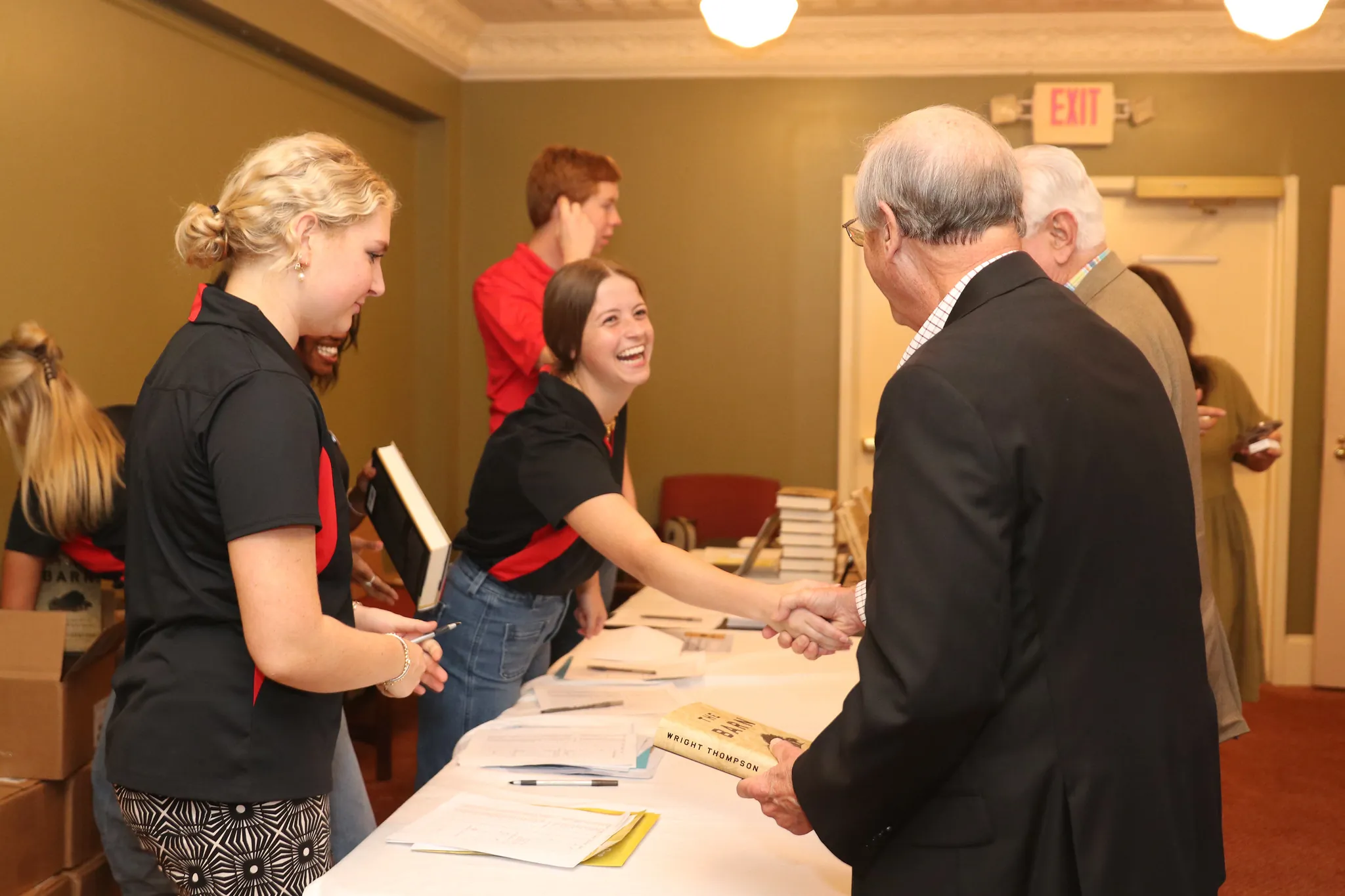Two young women, Grady College Ambassadors, smile and shake hands with two older men at a check-in desk in a warmly lit room. Books and papers are neatly arranged on the table.