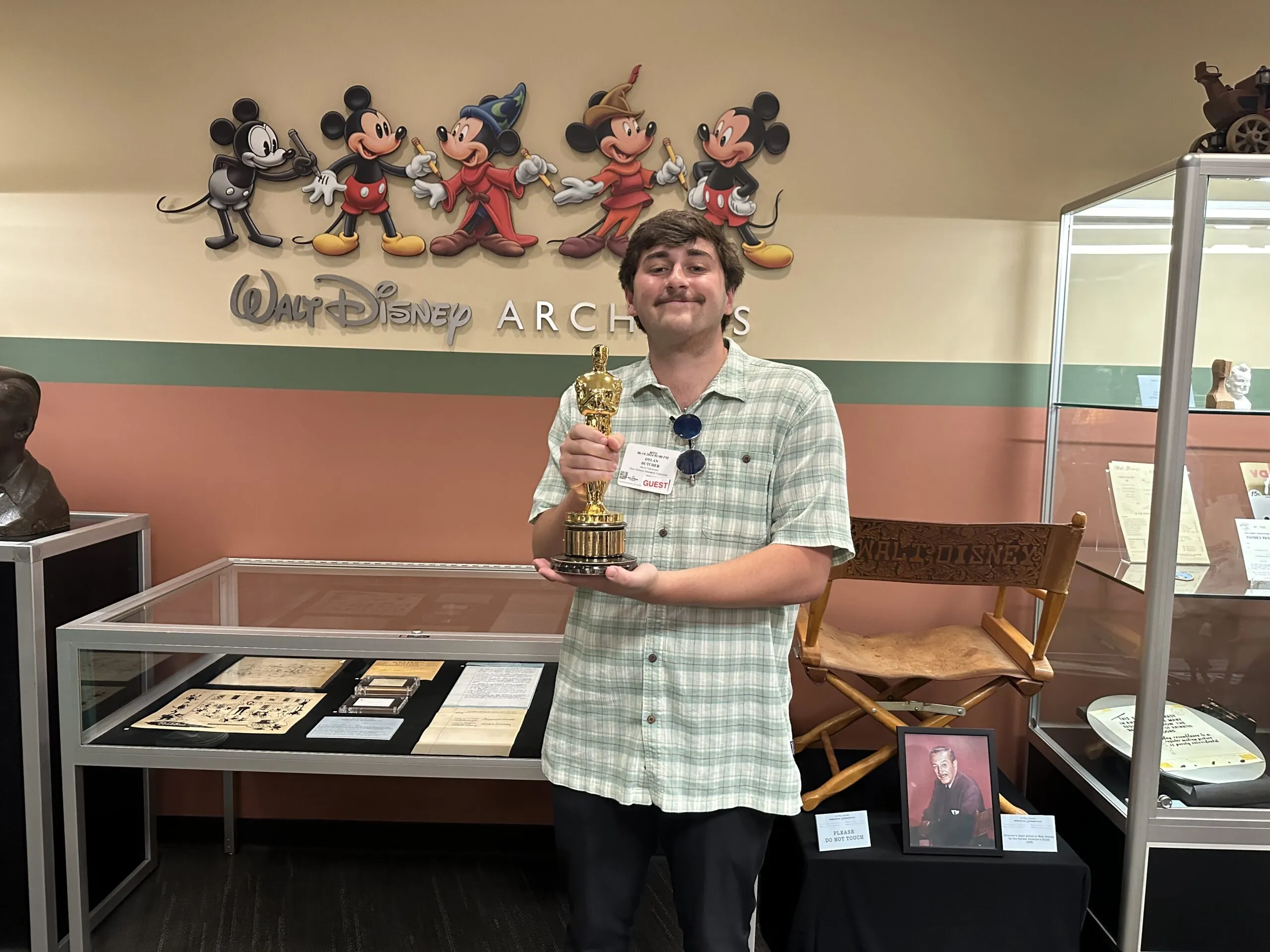 A person in a green plaid shirt, embodying tenacity, holds an Oscar trophy and smiles inside the Walt Disney Archives, with Mickey Mouse artwork and Disney memorabilia displayed behind him.