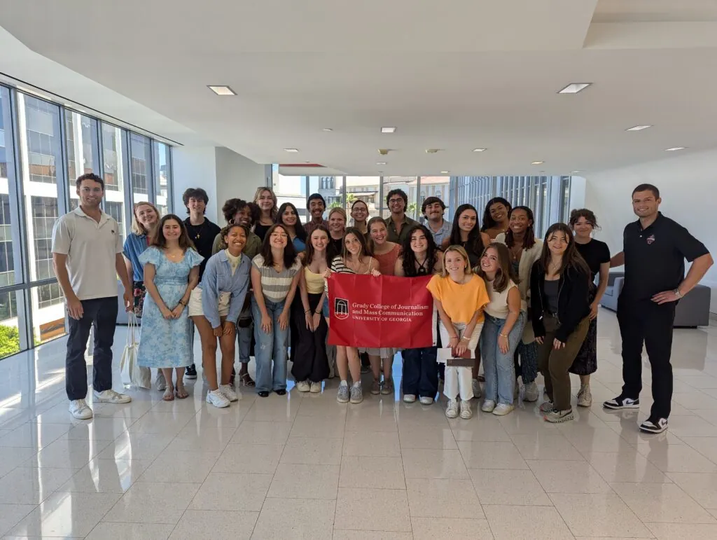 A group of students and two adults stand indoors, smiling and posing for a photo. Showcasing their tenacity, several people in the center proudly hold a red banner reading “Grady College of Journalism and Mass Communication, University of Georgia.”.