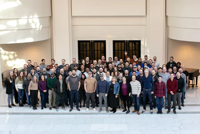 A large group of people, including Jason Russell, pose together for a photo in a spacious, well-lit indoor area with marble floors and tall walls, all facing the camera and smiling.