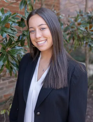 A young woman with straight brown hair, wearing a white blouse and a black blazer, smiles while standing outdoors in front of green leafy plants and a brick wall, representing the Grady College Ambassadors.