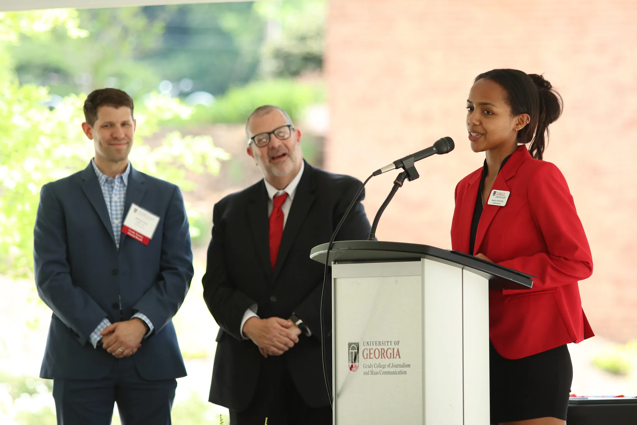A woman in a red blazer speaks at a podium labeled University of Georgia outdoors, while two men in suits—Grady College Ambassadors—stand nearby, listening and smiling.