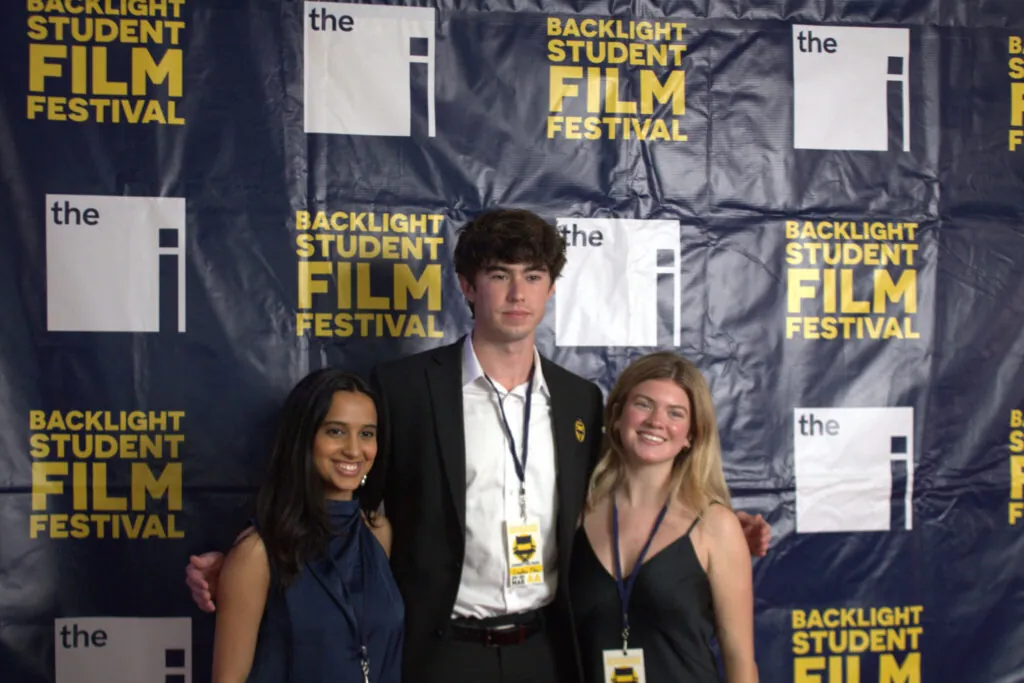 Three young adults pose and smile together in front of a backdrop that reads Backlight Student Film Festival, celebrating the Profiles of Tenacity. Two women flank a man in a suit, all wearing festival lanyards.