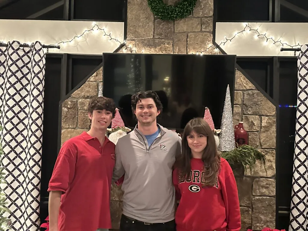 Three people stand smiling in front of a fireplace decorated with holiday wreaths, garlands, gnomes, and string lights. Two wear University of Georgia shirts; one wears a light gray pullover—true Profiles of Tenacity celebrating the season together.
