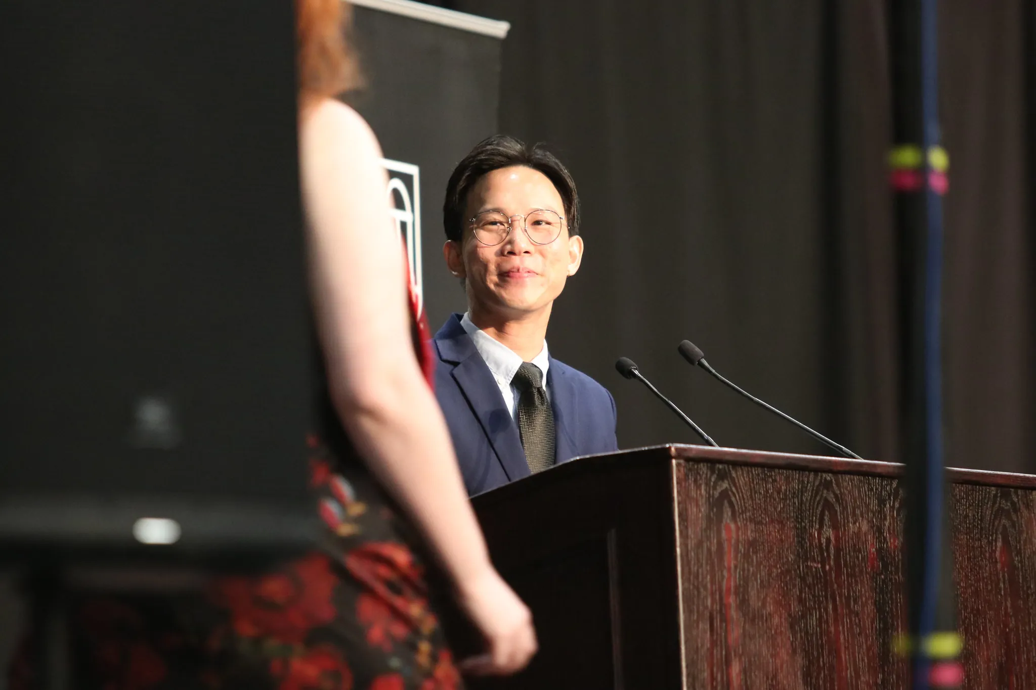 A man in a suit and glasses stands at a podium with two microphones during the Grady Convocation, smiling at a person in a floral dress who is facing him. The background is dark and there is a speaker in the foreground.