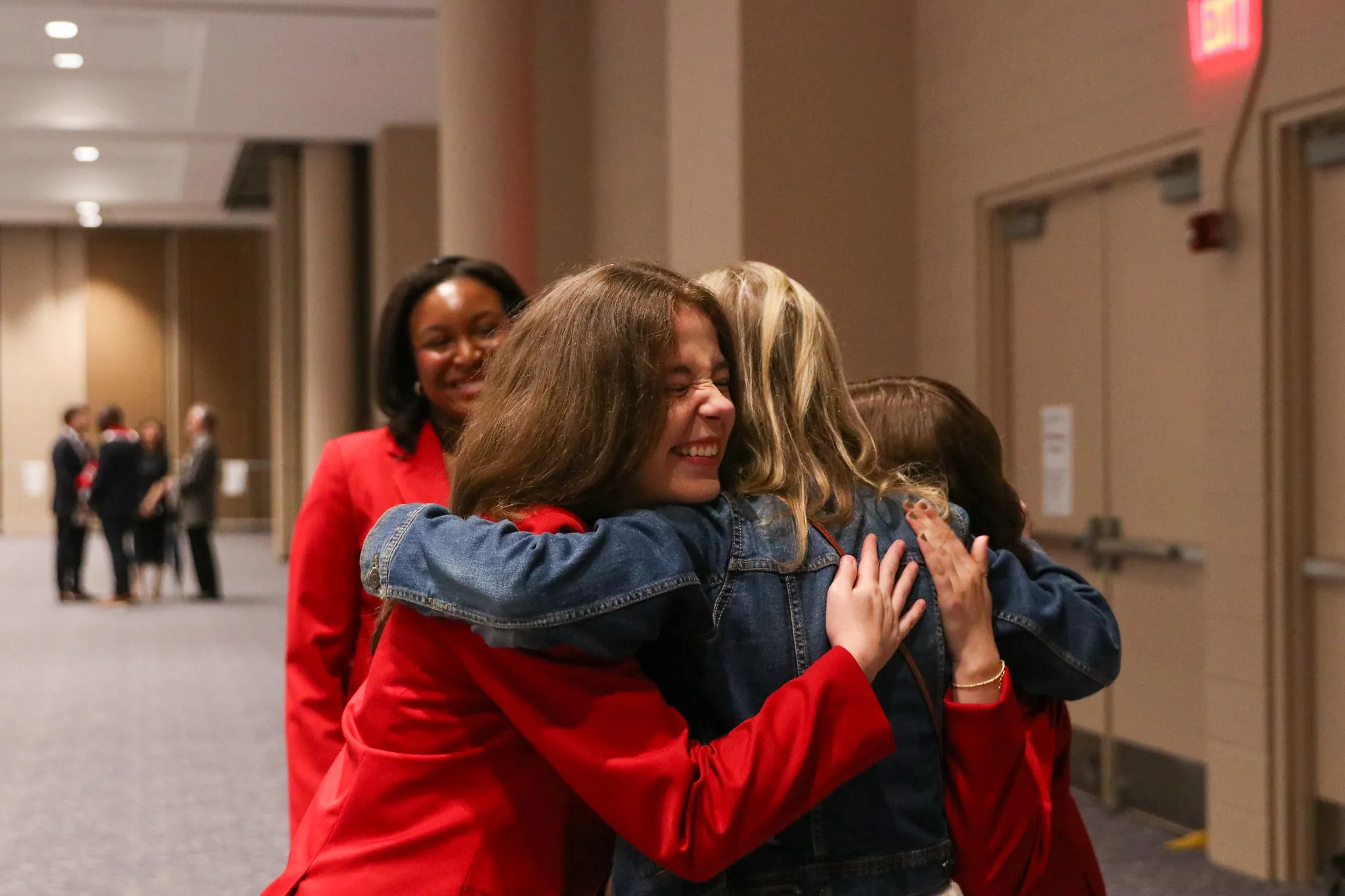 Three young women, two in red blazers and one in a denim jacket, hug each other and smile warmly in a hallway during Grady Convocation, while another woman in a red blazer stands nearby, also smiling.