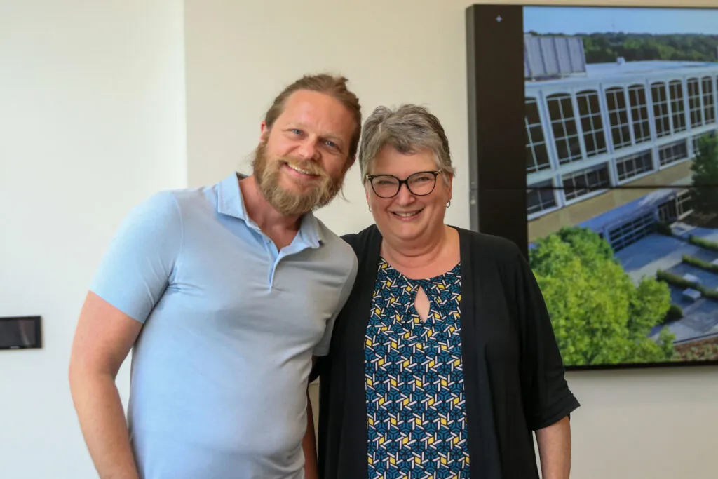 A man in a light blue shirt and a woman with glasses and gray hair stand smiling together indoors, celebrating as the 2025 faculty and staff award recipients announced artwork of a building hangs on the wall behind them.