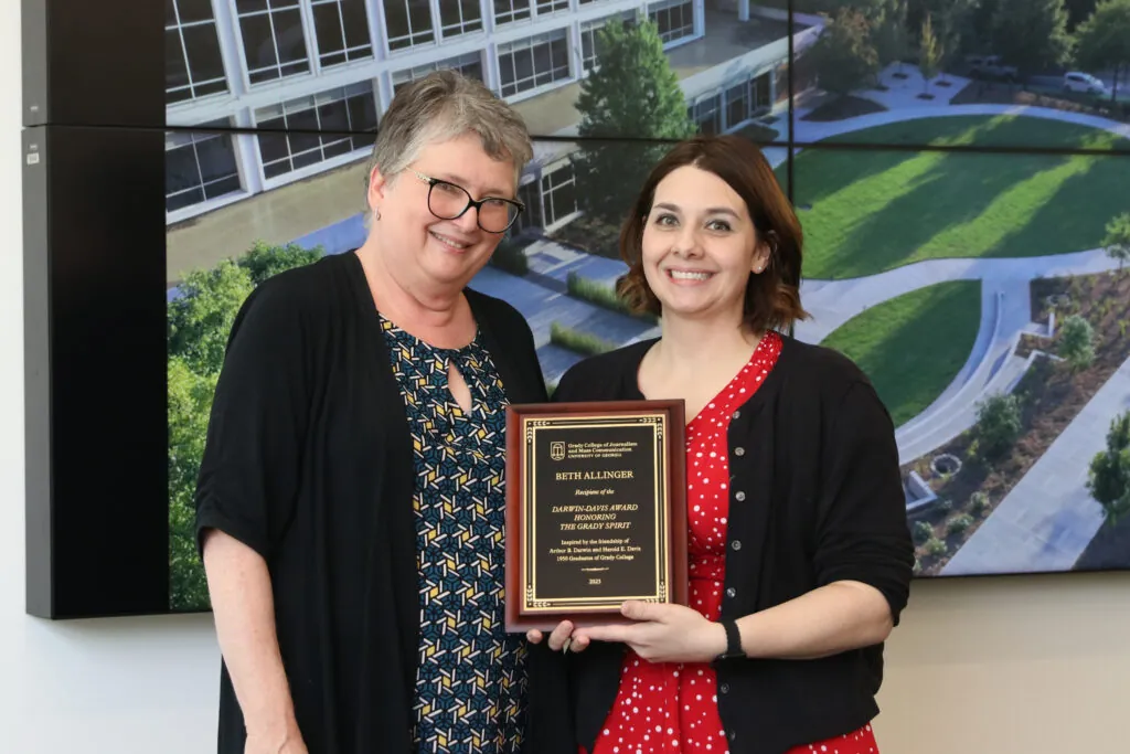 Two women stand together indoors, smiling at the camera. One holds a plaque celebrating the 2025 faculty and staff award recipients announced, while a large screen displays an aerial view of a campus in the background.
