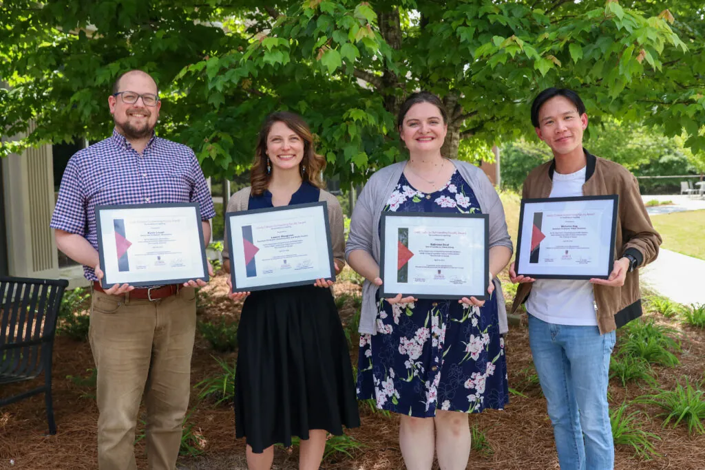 Four people stand outdoors in front of green trees, smiling and holding framed certificates. Dressed in business casual attire, they appear to be celebrating as the 2025 faculty and staff award recipients are announced.