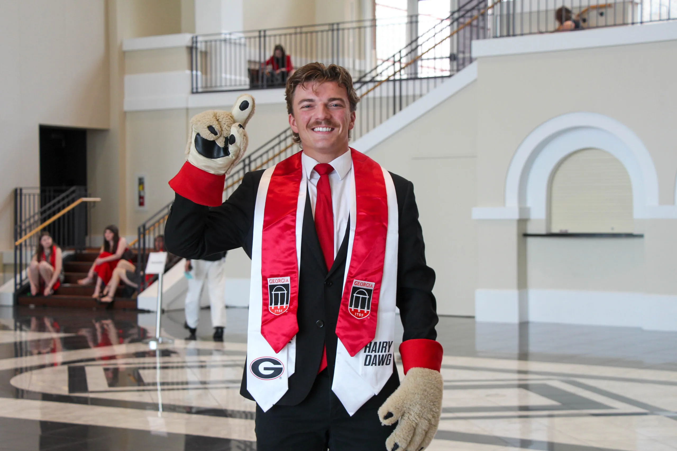 A smiling graduate in a black suit, red tie, and University of Georgia sash celebrates Grady Convocation indoors, wearing large mascot gloves and holding up one finger in a triumphant gesture.