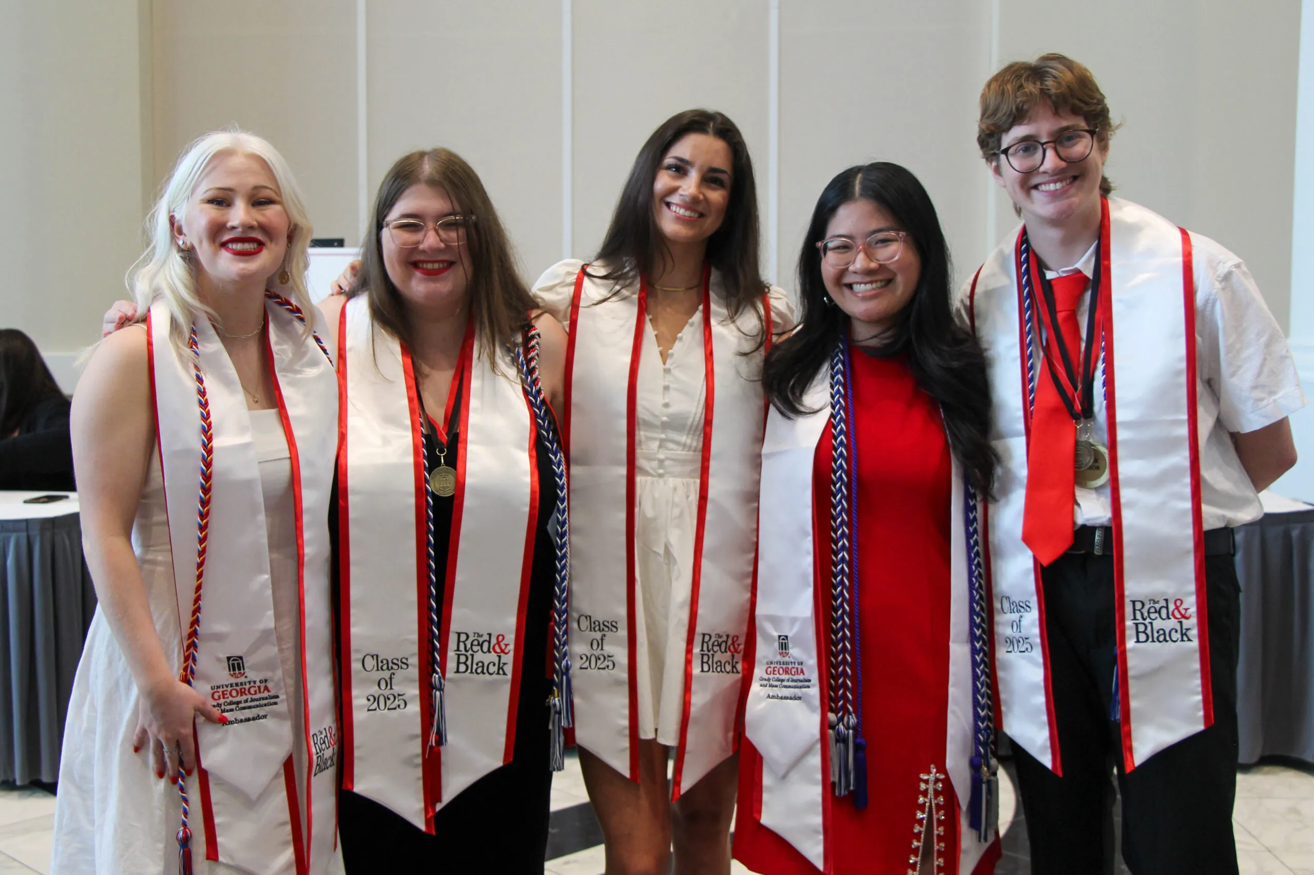 Five smiling graduates wearing white stoles, some with medals, stand together at Grady Convocation. Four wear red, white, and black cords. Dressed in white or red outfits, they celebrate the Class of 2023 indoors.