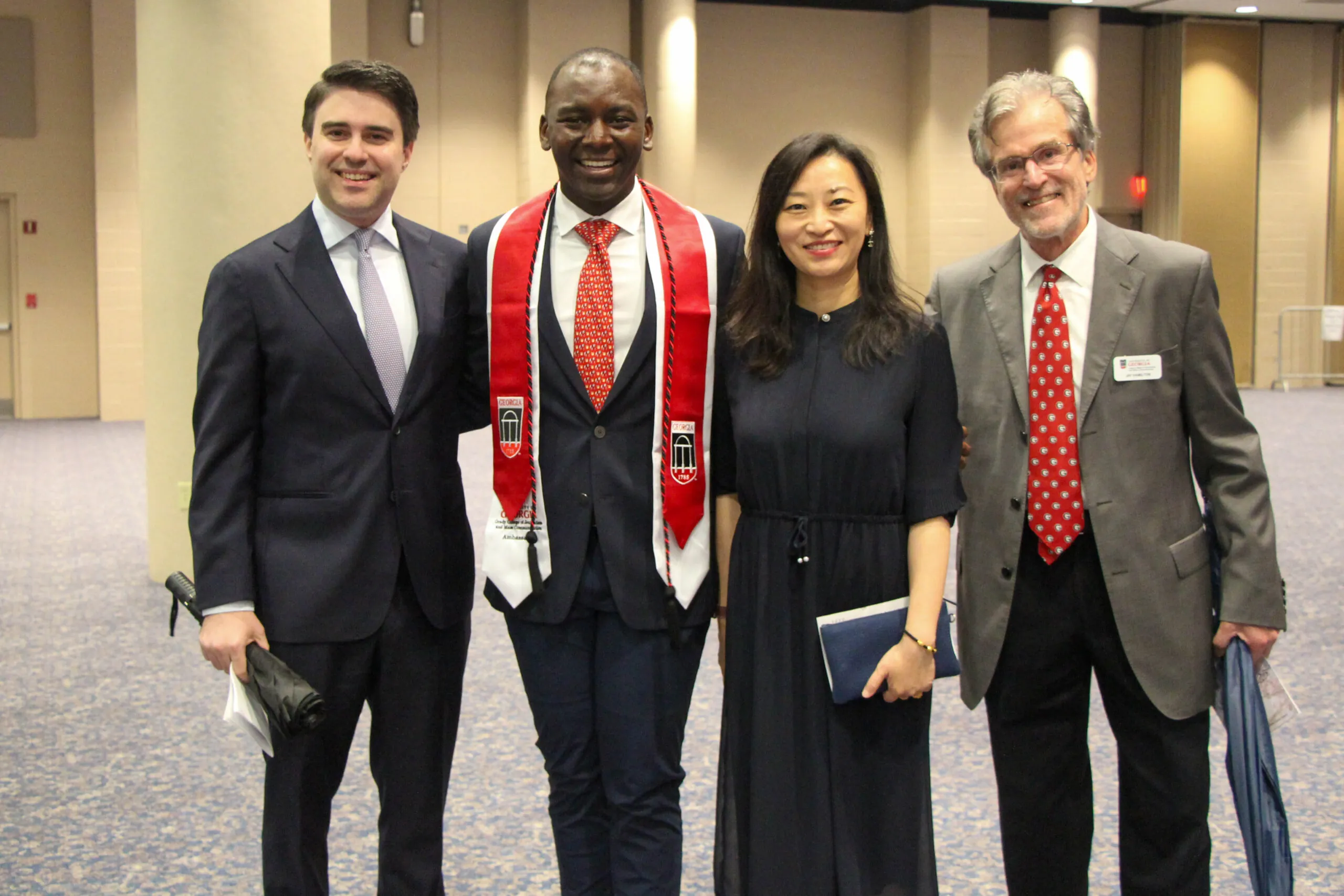 Four adults stand together indoors, smiling at the camera. One man in the center wears a Grady Convocation stole and suit; the others are dressed in formal attire. They appear to be at an event or ceremony in a large room.