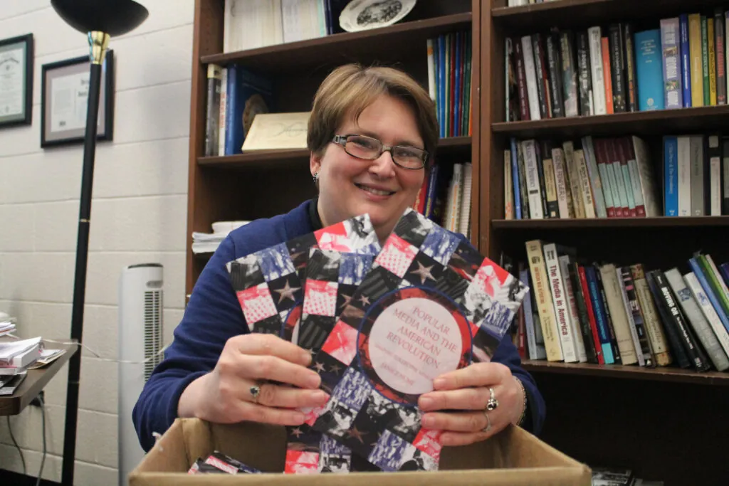 Janice Hume sits in her office and holds copies of her new book "Popular Media and the American Revolution."